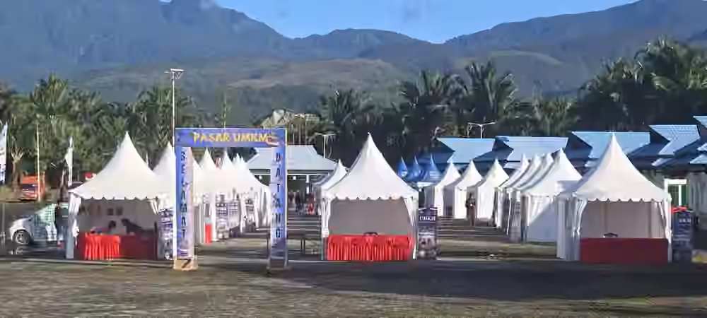 Deretan tenda booth event outdoor dengan gerbang Pasar UMKM dan latar pegunungan Papua.
