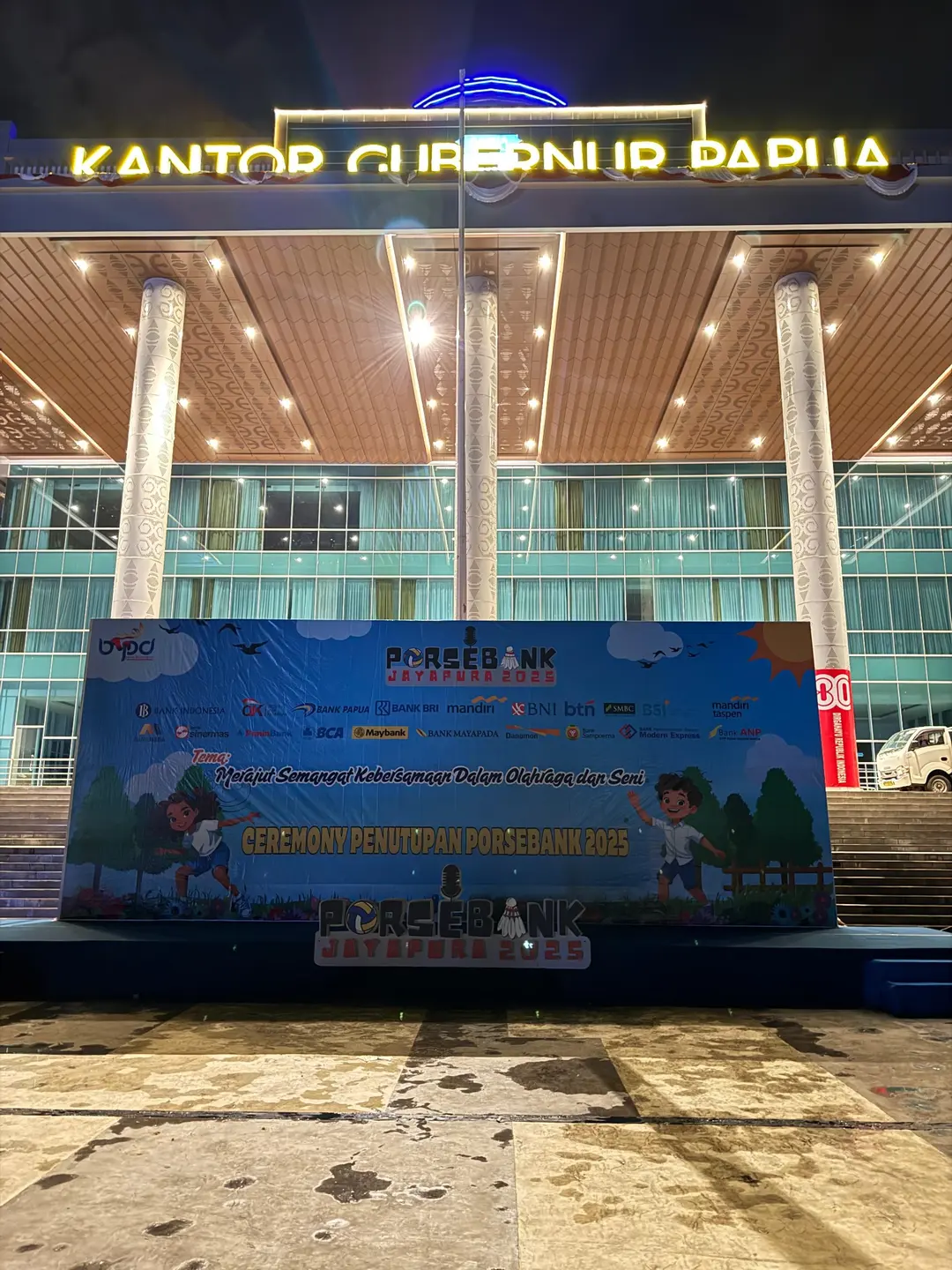 Panggung malam hari dengan backdrop acara dan pencahayaan gedung.