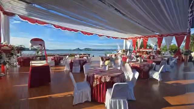 Tenda event outdoor dengan meja bundar, kursi, dekorasi merah-putih, dan pemandangan laut.