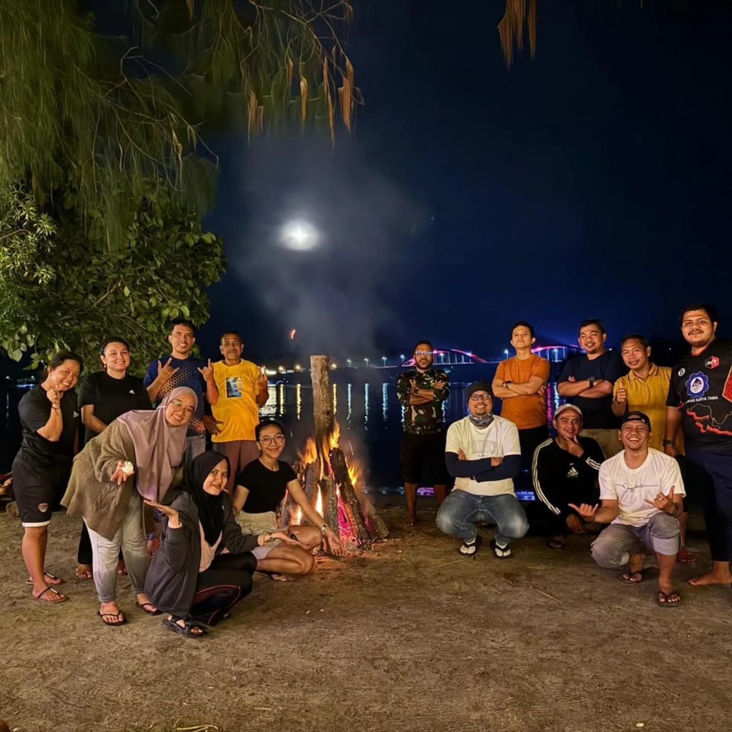 Rombongan tamu berfoto di sekitar api unggun malam hari, Jembatan Merah menyala dan bulan purnama di latar belakang